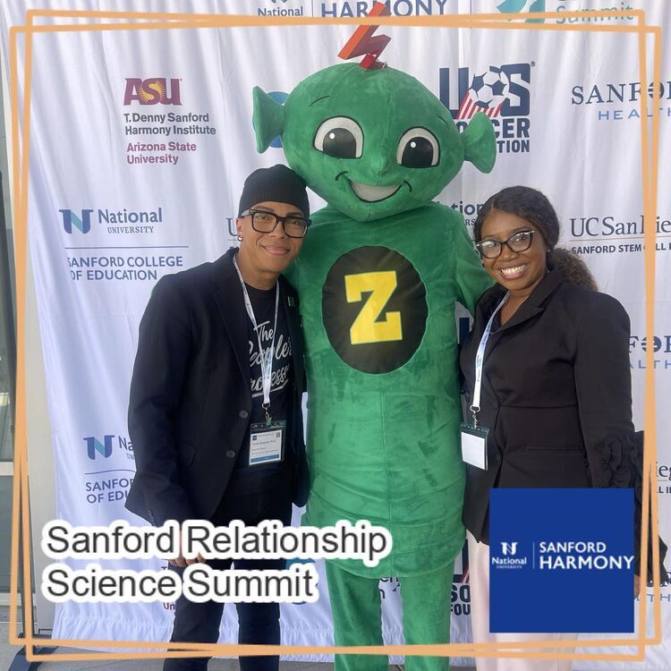 Two attendees pose with the Sanford Harmony mascot in front of the summit sponsor backdrop