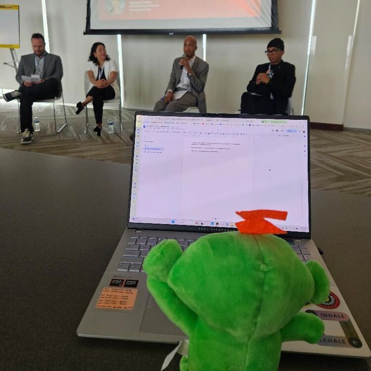 A Sanford Harmony mascot plush sits beside a laptop as four panelists speak on stage