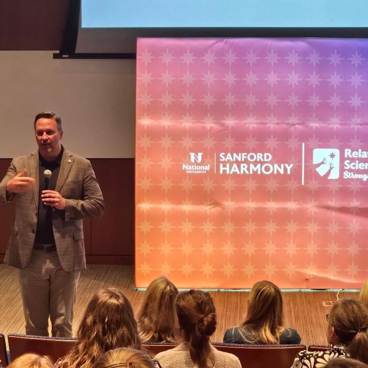 A speaker addresses the audience in front of the Sanford Harmony stage backdrop